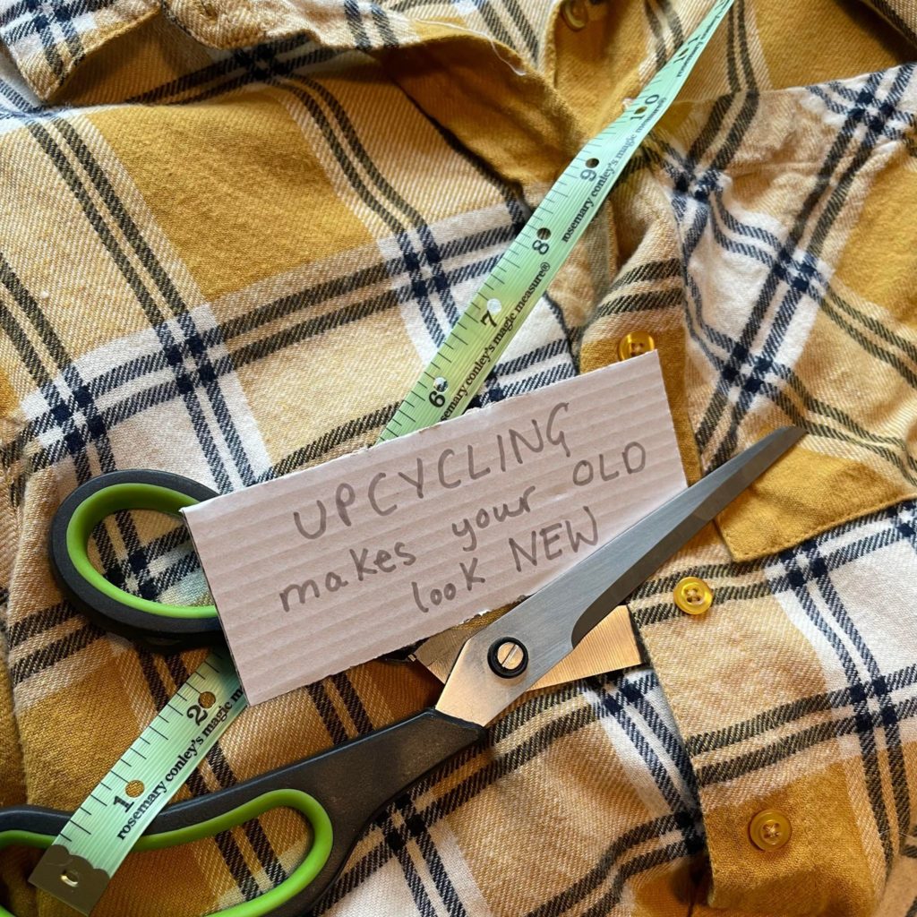 Scissors and tape run across a yellow flannel shirt. Sign reads: Upcycling makes your old look new.
