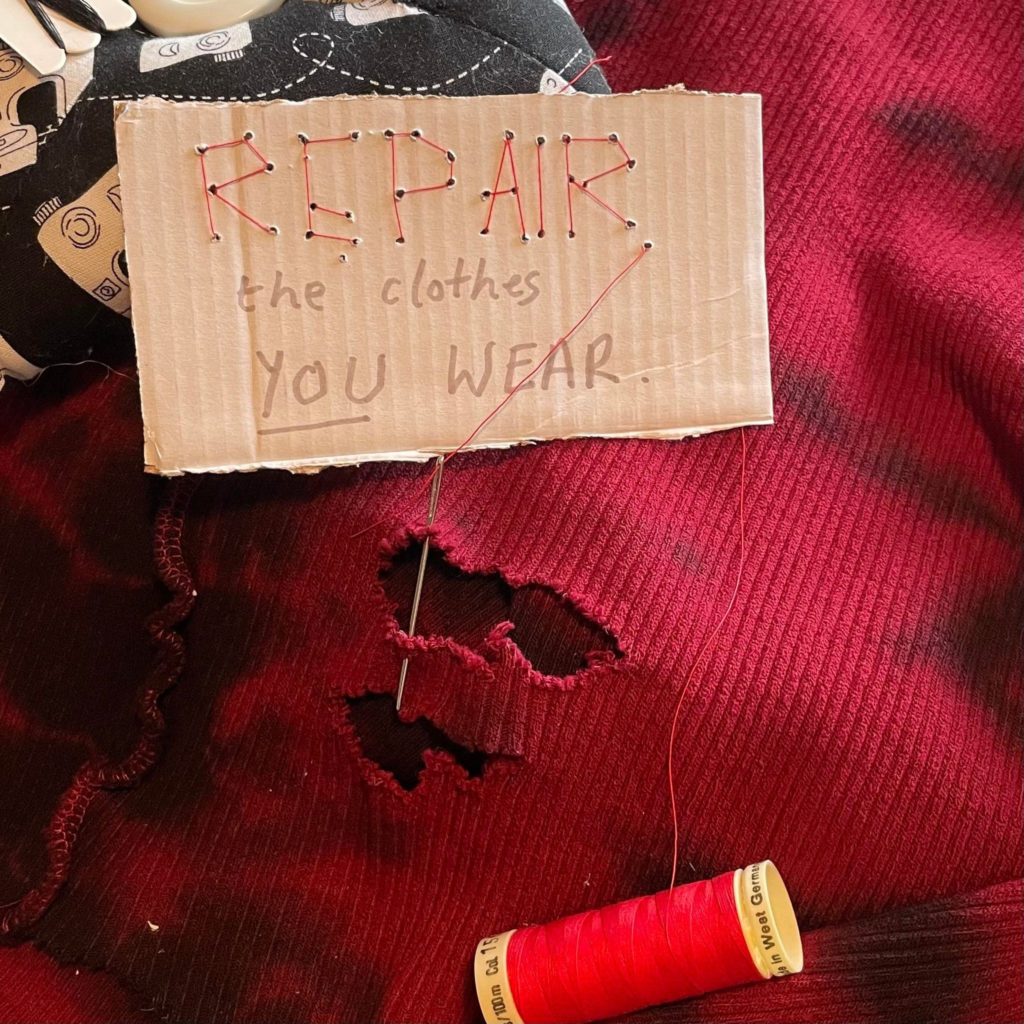 Red thread on a needle dewing up a hole in a red garment. Sign reads: Repair the clothes you wear