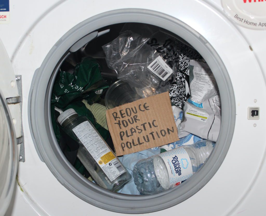 Image of a washing machine, containing miscellaneous items made of plastic, including plastic bottles and plastic bags. In the centre of the image is a piece of cardboard with 'Reduce your plastic pollution' written on it
