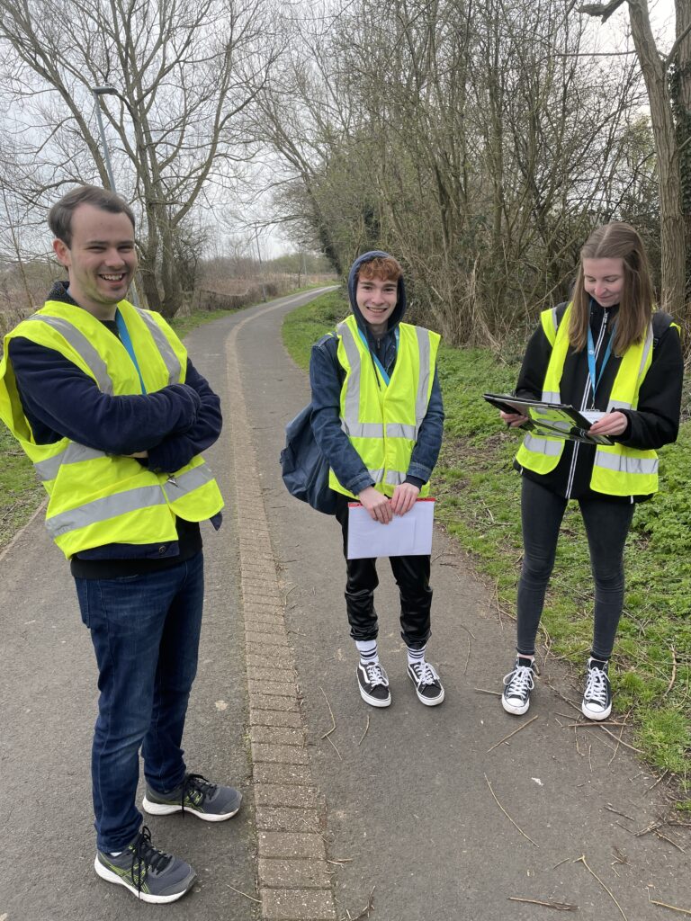 Three students wearing Hi Visibility jackets and carrying clip boards